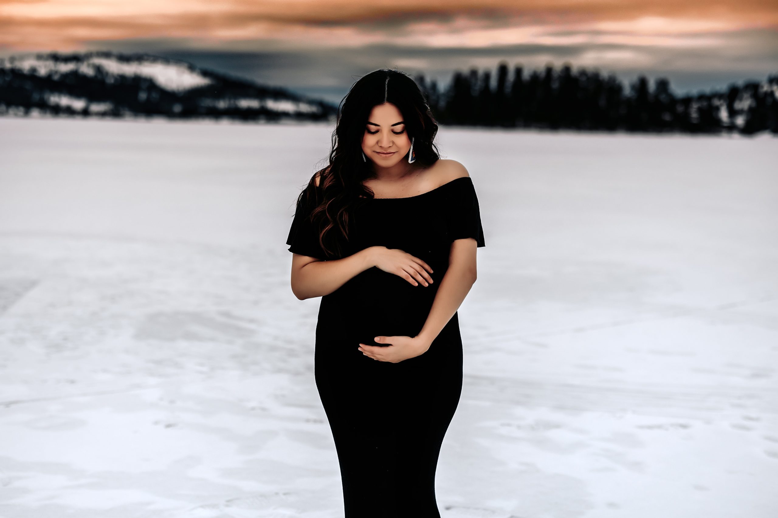 pregnant woman cradling her stomach on a frozen lake in kalispell, mt with an orange sunset behind her