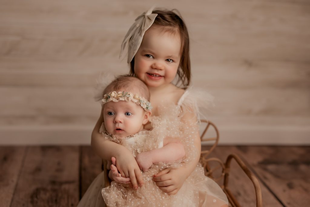 sibling babies dressed in neutral dresses sit together and smile at the camera
