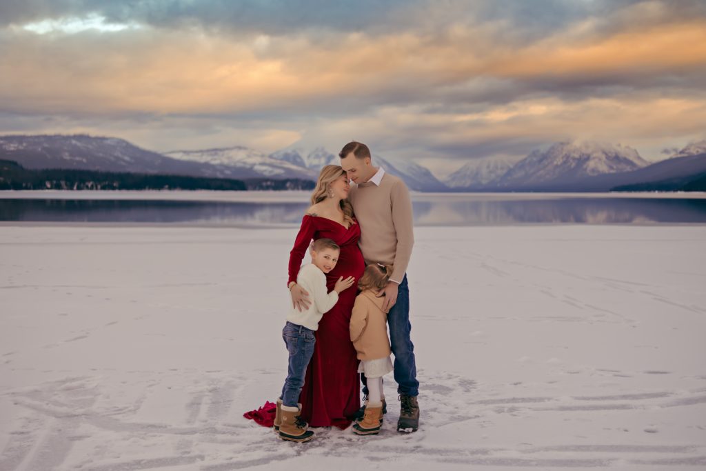 A FAMILY STANDS IN FRONT OF THE MOUNTAINS ON A FROZEN LAKE IN GLACIER NATIONAL PARK