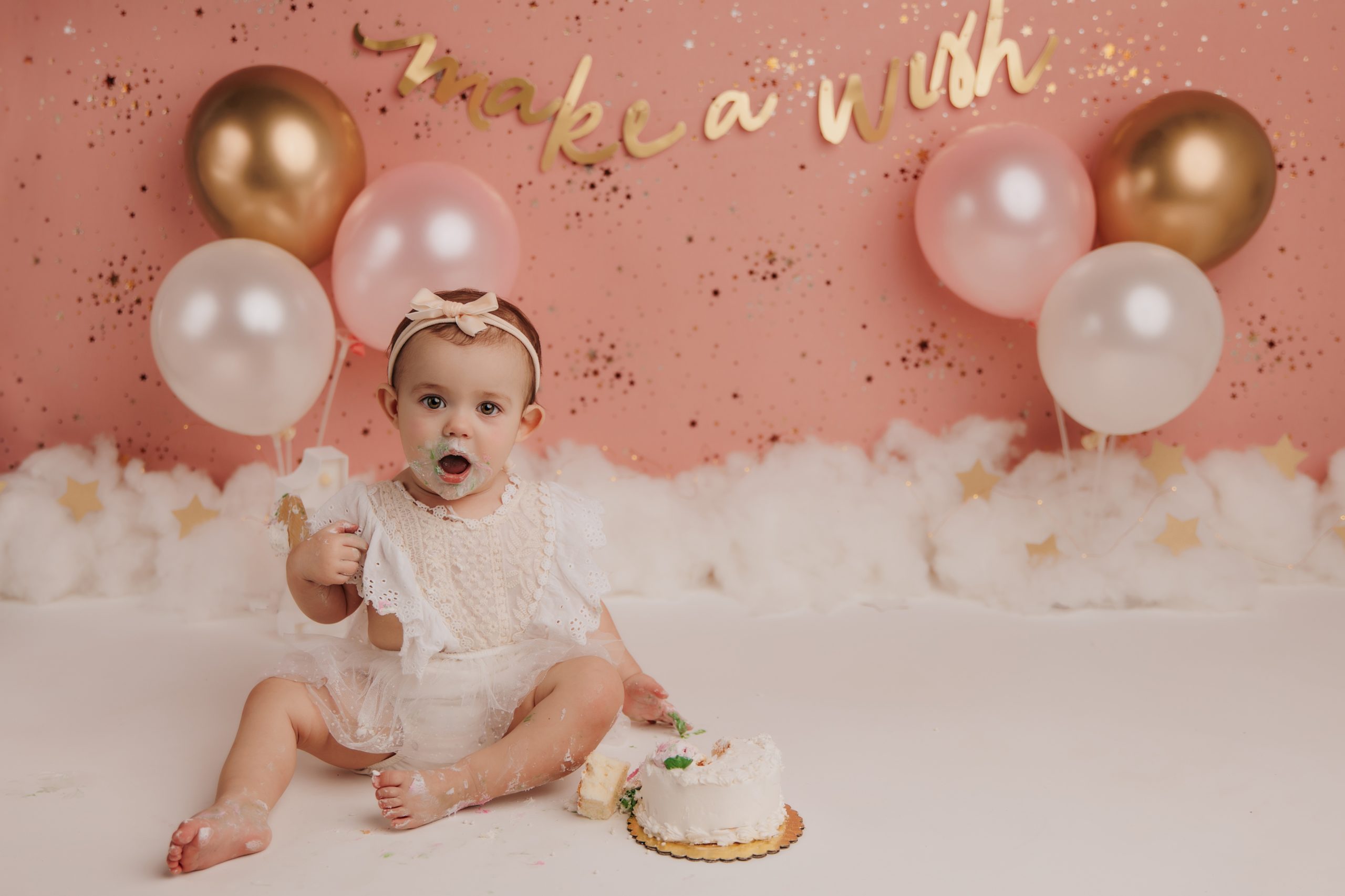 baby girl sits in front of pink and gold backdrop with cake and look of surprise on her face