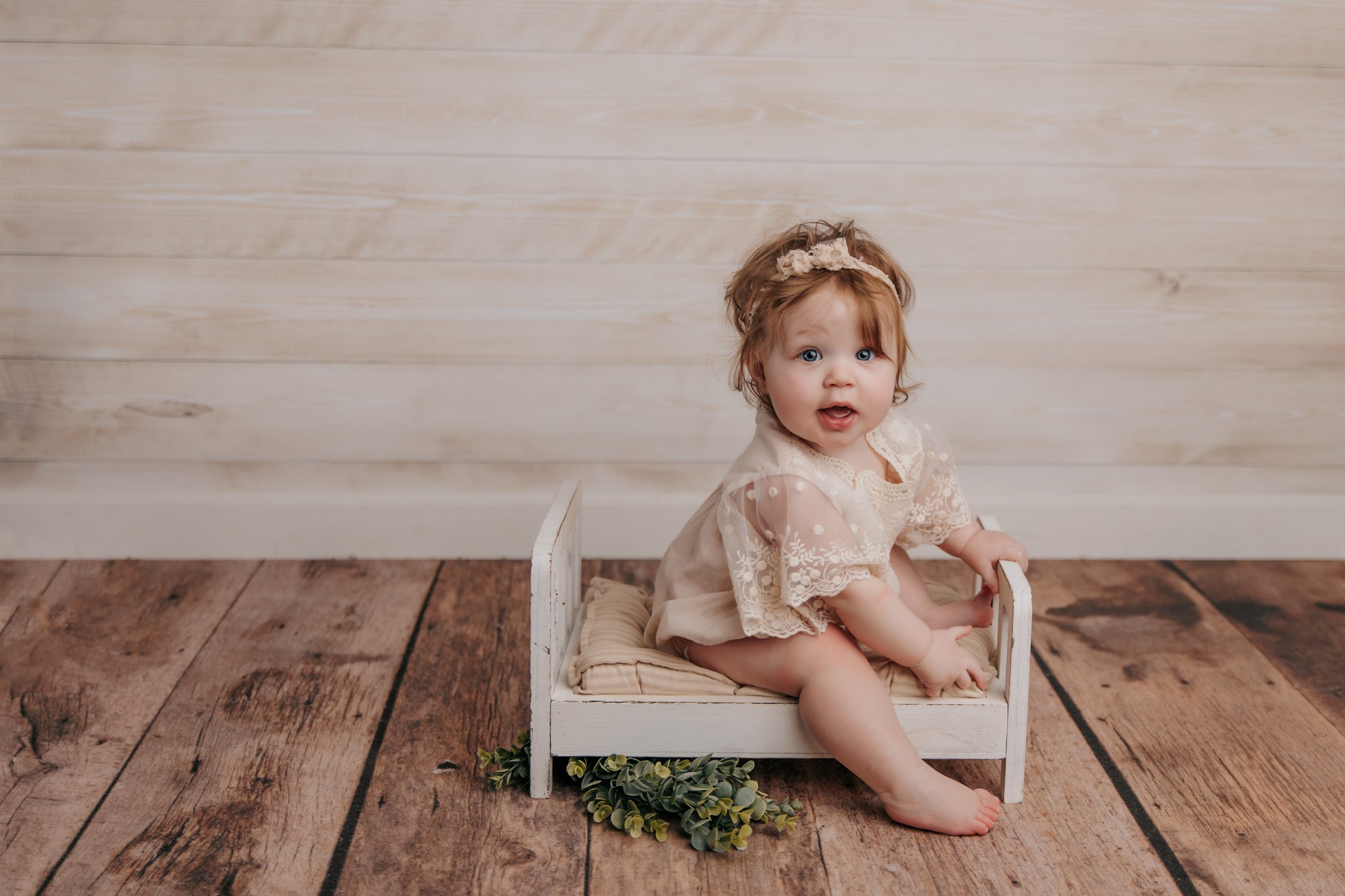 six month baby girl sits on small white bed in front of rustic wood backdrop